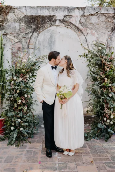 Wedding kiss portrait as the bride and groom kiss beneath an arched stone niche, her bouquet contrasting with the brick floor and garden greenery.