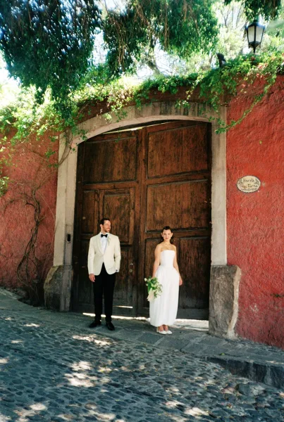 Couple portrait, an outdoor wedding portrait: bride holds a bridal bouquet beside the groom by a red stucco wall and ivy-clad gate.