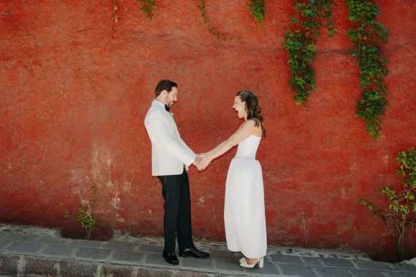 Wedding first look with a couple holding hands, smiling face to face against a red stucco wall with vines; groom in a white tuxedo jacket.