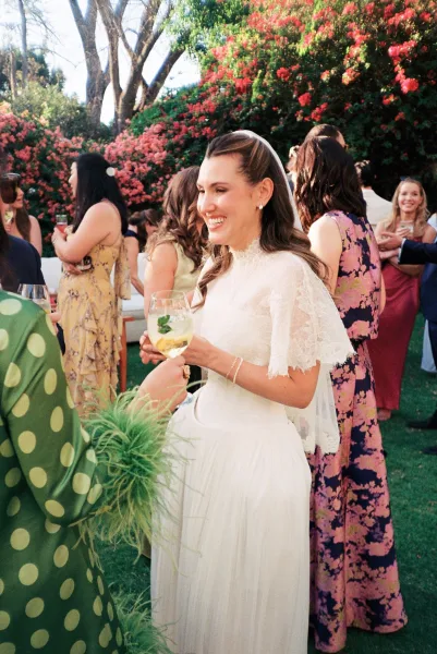 Cocktail hour moment as a bride with cocktail mingles with friends in a sunlit garden, her lace capelet catching light by flowering bushes.
