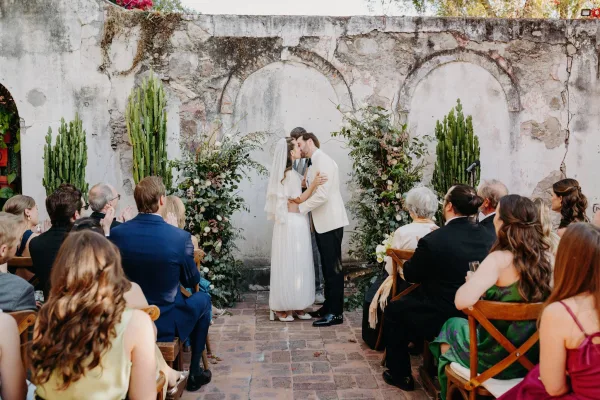 Ceremony kiss moment at an outdoor courtyard ceremony, framed by floral pillars against a weathered stone wall with arched niches.