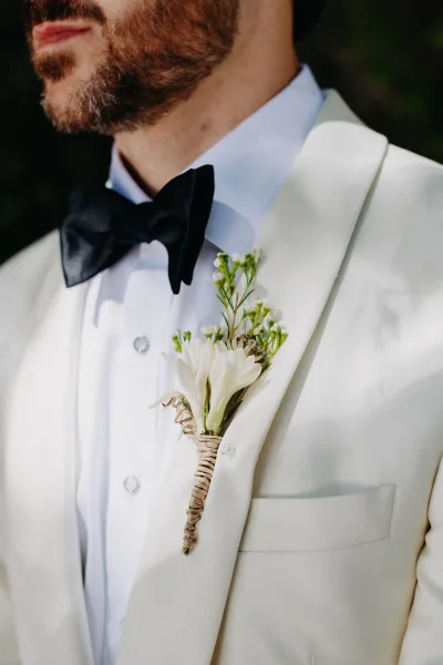 Groom boutonniere—a white floral boutonniere with fresh greenery and buds, pinned to a white dinner jacket.