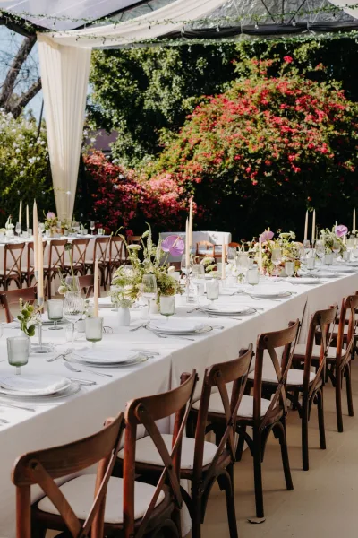 Reception tablescape beneath a clear tent canopy, lined with taper candles amid garden trees and flowering bushes in the background.