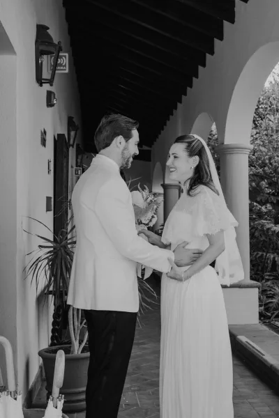 Couple portrait of a bride and groom smiling, embracing under an arched hallway with columns, her lace bridal cape draping softly.
