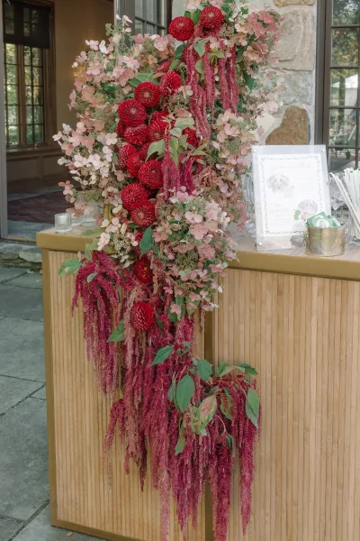 Wedding welcome sign on an acrylic frame with cascading florals and hanging amaranthus beside a wooden bar against a stone wall backdrop