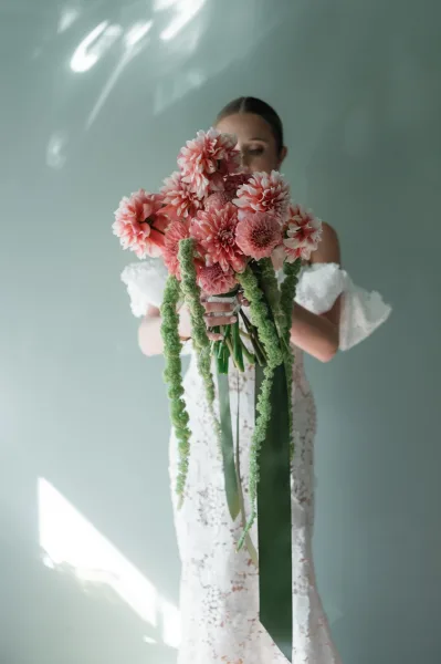 Bridal bouquet with pink dahlia bouquet and cascading green amaranthus, tied with a long ribbon, held against a sage green wall in window light