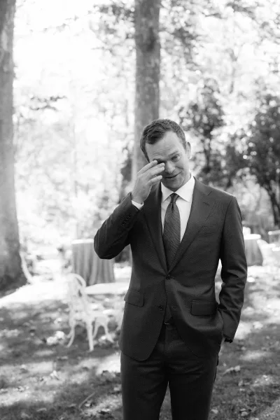 Groom portrait of a groom wiping tears in a two-piece suit and necktie, standing in a garden with trees and dappled sunlight