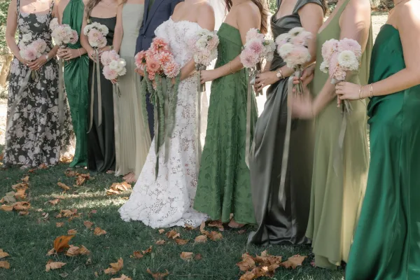 Wedding party portrait of bride with bridesmaids in green dresses and groom in navy suit, holding dahlia bouquets with ribbon wraps on a lawn with trees