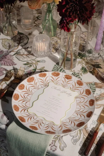 Reception tablescape with wedding place setting featuring a menu on the dinner plate, dahlias in bud vases, and candlelit brass flatware under soft lighting
