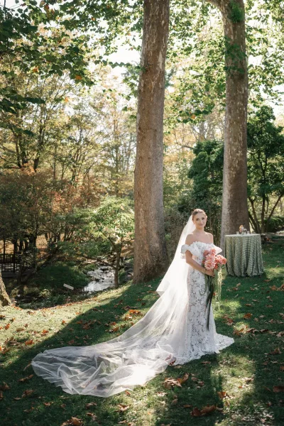 Bridal portrait of a bride holding bouquet of dahlias with cascading greenery, wearing a lace off-shoulder dress and long veil in woodland trees