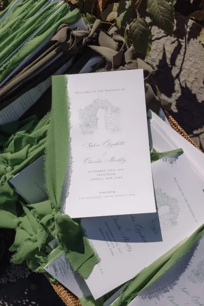 Wedding invitations in a wedding invitation suite with deckle edge paper and green ribbon bows, arranged in a wicker basket on stone in sun shadows
