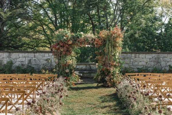 Ceremony setup for an outdoor wedding ceremony with a floral arch wedding, wooden crossback chairs, and a stone wall backdrop on grass