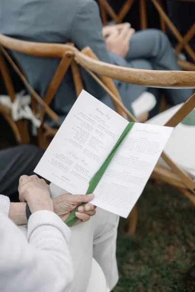 Wedding program with green ribbon tied around a printed ceremony booklet, held by a guest among wooden folding chairs on a lawn