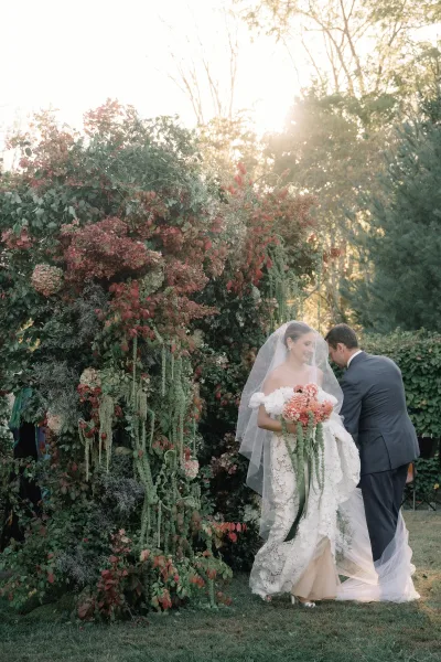Couple portrait of bride and groom portrait with her veil and bouquet before a floral wall, on a sunlit garden lawn with trees