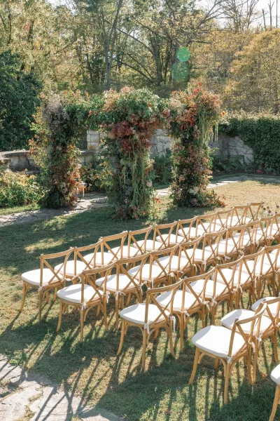 Outdoor ceremony setup with natural wood crossback chairs and white cushions facing a floral arch on stone pavers, framed by ivy wall