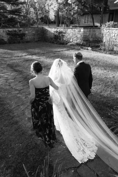Processional moment as the bride walking to ceremony in a long cathedral veil, seen from behind on a stone path by garden lawn