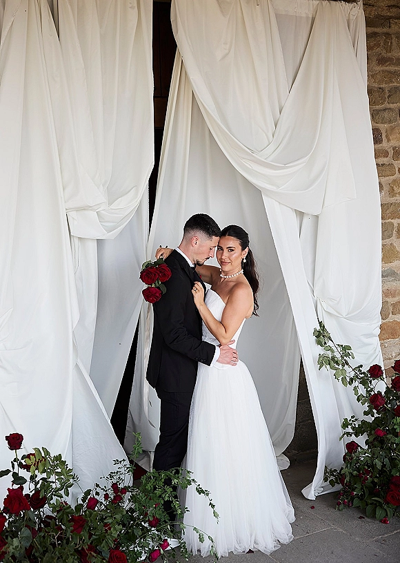 Couple portrait of bride and groom embrace, groom holding bouquet of red roses, in a doorway with white draped fabric backdrop behind them