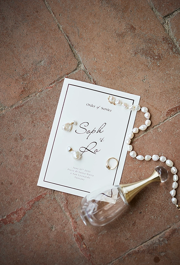 Wedding invitation suite wedding stationery flat lay with program card, pearl jewelry, engagement ring, and champagne coupe on terracotta tile floor