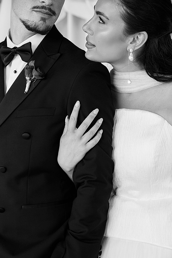Couple portrait in a black and white wedding portrait, bride holding groom’s arm in side profile, tuxedo with boutonniere and strapless dress indoors
