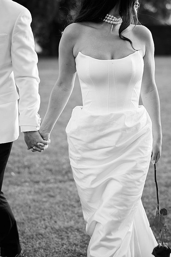 Couple portrait of bride and groom holding hands, showcasing pearl choker, strapless dress, rings, and rose stem on a grassy lawn