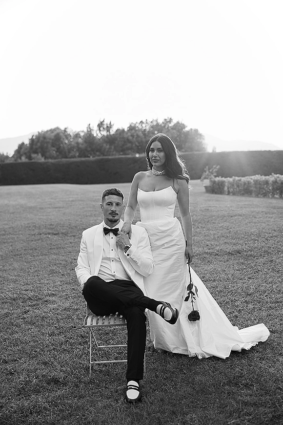 Couple portrait in a black and white wedding portrait style, bride standing with a black rose beside seated groom in white tux on a lawn