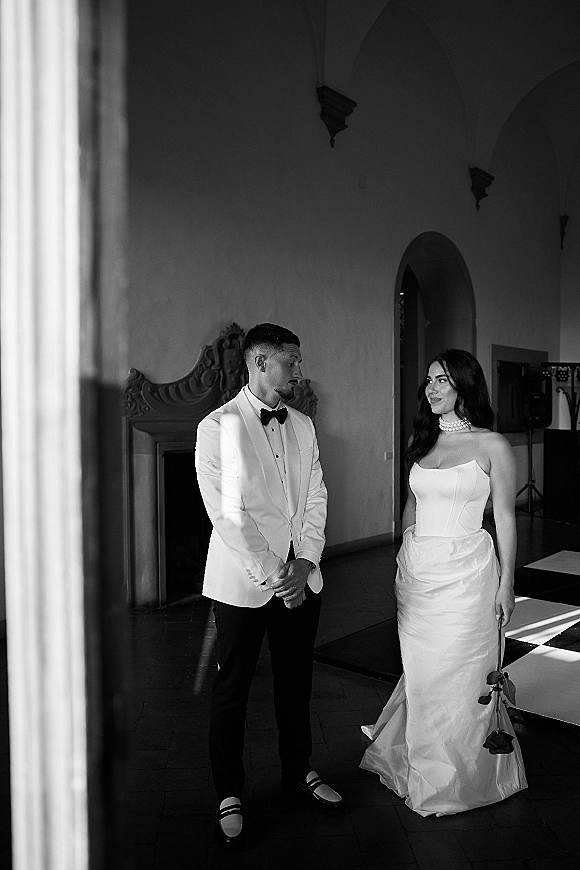 Couple portrait in black and white, bride in strapless gown with pearl choker holding a single rose, groom in white tuxedo jacket in arched hallway