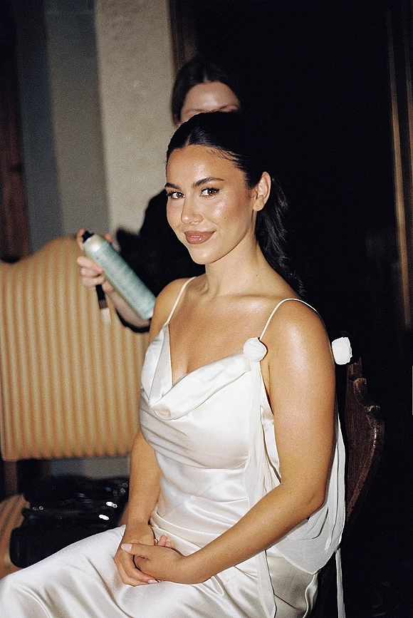 Bridal portrait of a bride getting ready in a satin spaghetti-strap dress, seated as stylist sprays her low ponytail in an indoor room