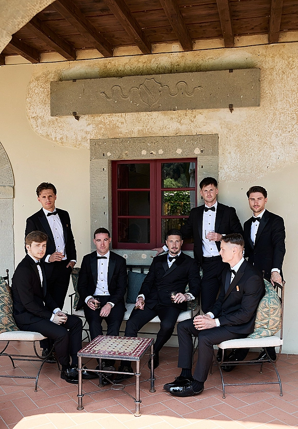 Groomsmen portrait in black tuxedos and bow ties holding whiskey glasses, posed on patio lounge chairs by a stucco wall and window