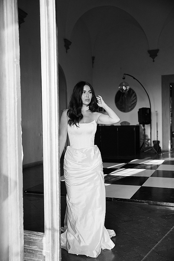 Bridal portrait in a strapless wedding dress as the bride adjusts her hair by an arched doorway with window light and checkered floor.