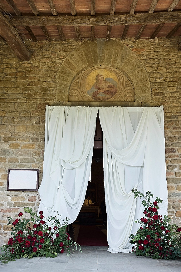 Ceremony entrance decor with white drapery framing an arched church doorway, red rose and greenery arrangements against stone walls and fresco mural