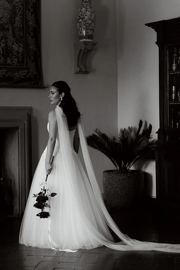 Bridal portrait of a bride looking back in a long veil and wedding dress, holding a rose bouquet in an indoor hallway with tapestry and palm