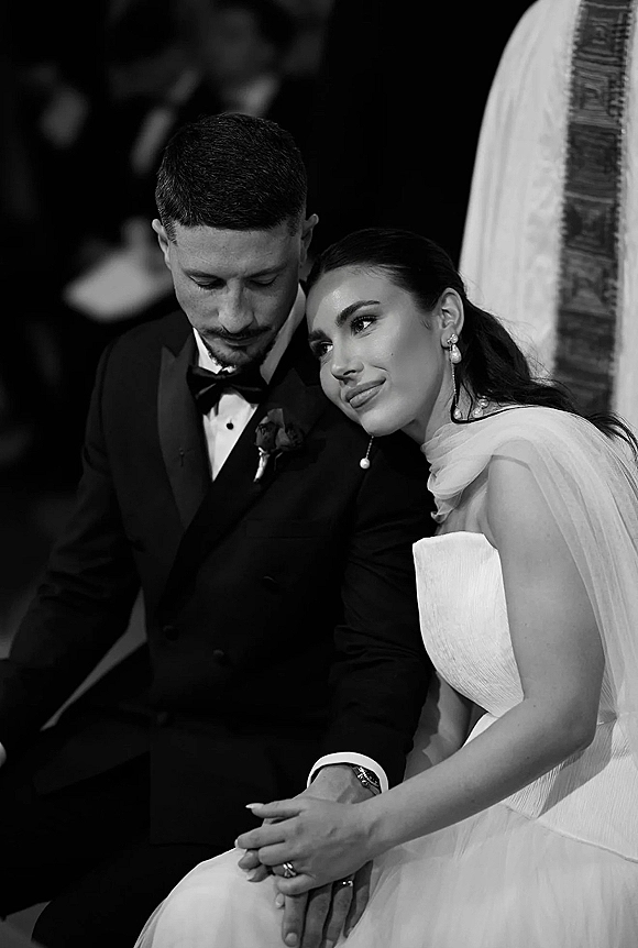 Couple portrait in a black and white wedding portrait, bride leaning on groom as they sit in an indoor ceremony with blurred guests behind