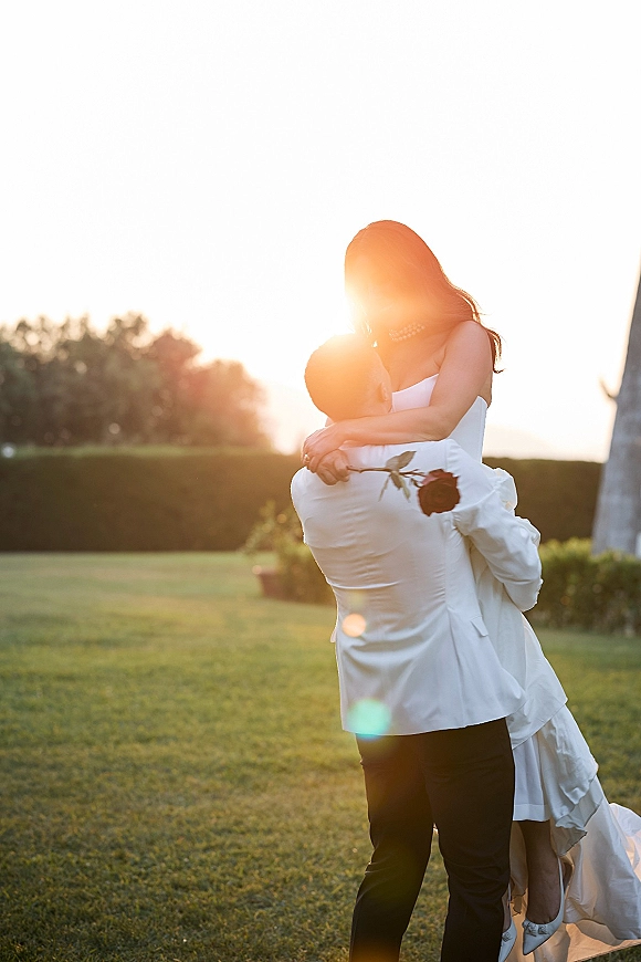 Wedding kiss in a dip as groom lifts bride in strapless dress and veil, holding a dark rose on a lawn at sunset near a stone building