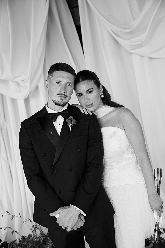 Couple portrait in a black and white wedding portrait, bride leaning on groom in tuxedo and strapless dress before draped curtains backdrop