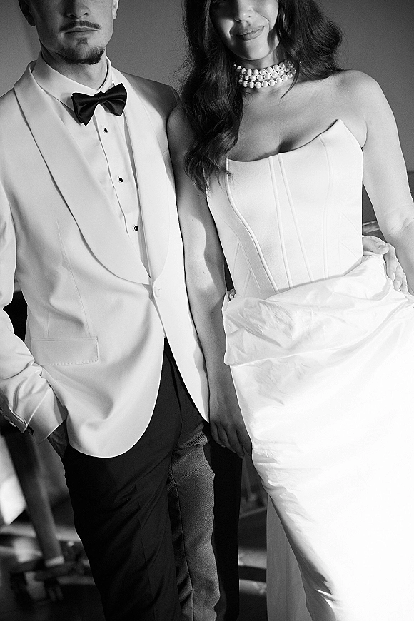 Couple portrait in black and white wedding portrait style, bride in strapless dress with pearl choker beside groom in white blazer and bow tie by window light