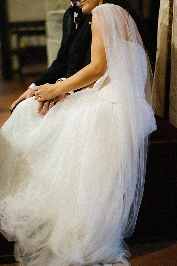 Couple portrait of bride sitting on groom’s lap, veil draped over strapless tulle dress as their ringed hands rest in warm light indoors