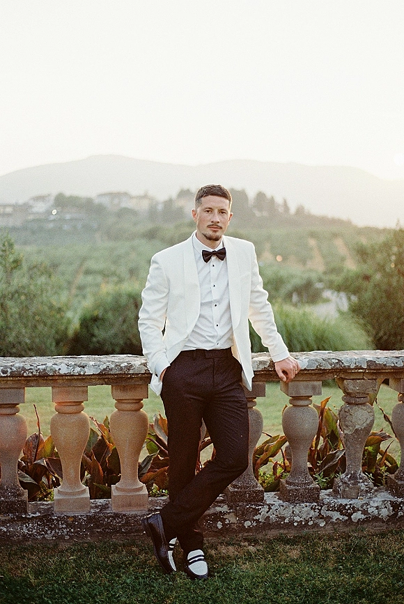 Groom portrait in a white tuxedo jacket with black bow tie, leaning on a stone balustrade on a terrace overlooking garden hills