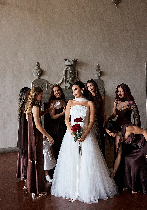 Bride with bridesmaids helping bride adjust her strapless gown and veil, holding a red rose bouquet against an ornate plaster wall indoors