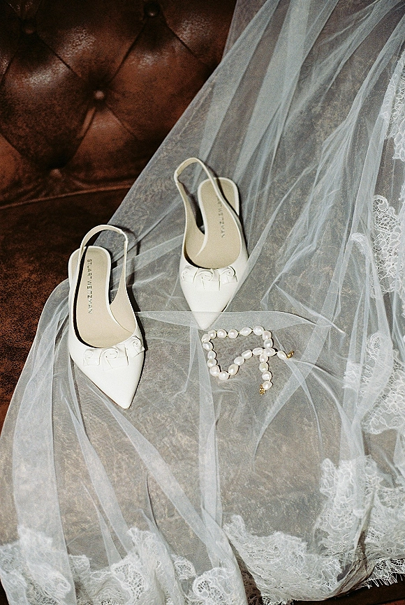 Bridal shoes, white slingback wedding heels with bow detail beside a pearl bracelet on a tufted leather couch with tulle veil backdrop