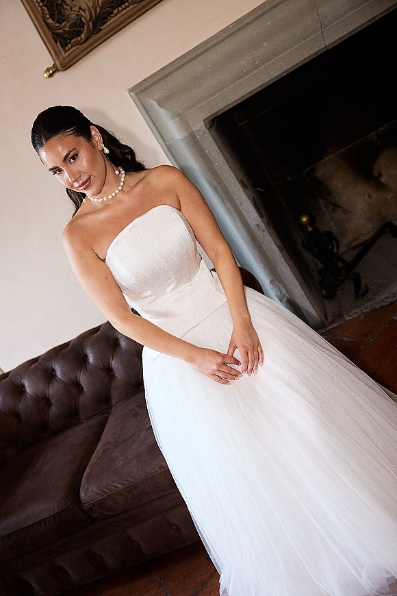 Bridal portrait of a bride in a strapless wedding dress with pearl necklace, leaning by a leather sofa near a fireplace indoors