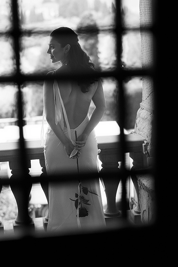 Bridal portrait in black and white of a bride from behind in a low back wedding dress, holding a rose on a stone balcony by water view