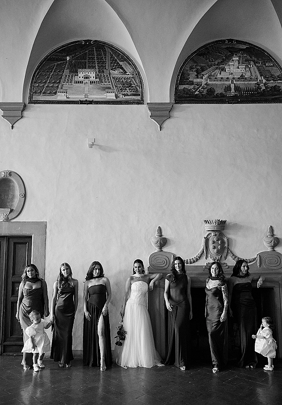 Bridesmaids portrait with bride and wedding dress, bridesmaids in black dresses holding bouquets in an arched hall with mural panels