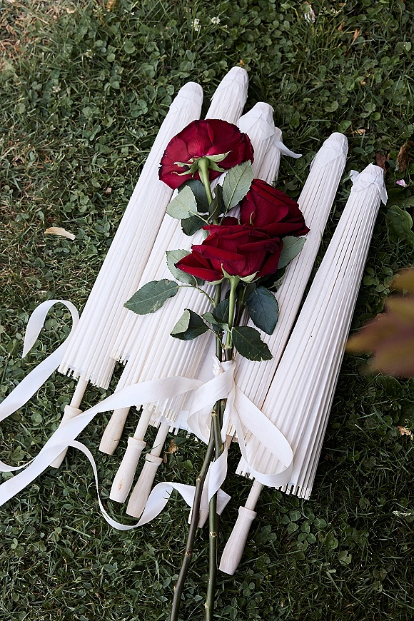 Wedding ceremony fans with paper hand fans, wood handles, and white ribbon tied to red rose and greenery stems on clover grass