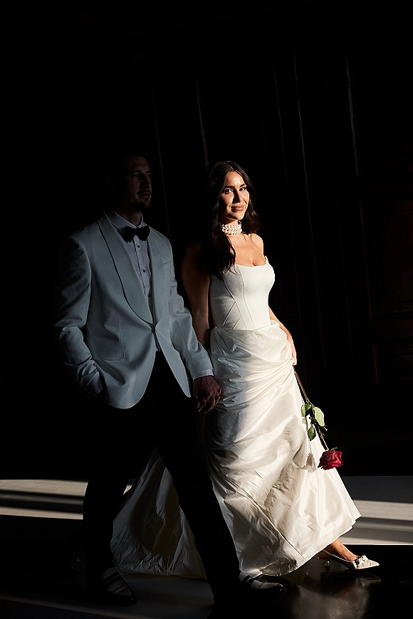Couple portrait of bride and groom holding hands, bride in strapless satin dress with red rose bouquet against dark wall shadows