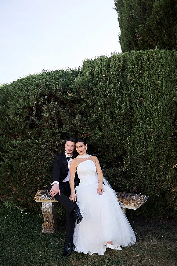 Couple portrait of bride and groom sitting on a stone bench, she leans on him in a strapless dress and veil by green hedges