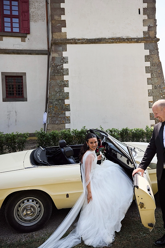 Wedding exit as groom opens convertible car door for bride in veil and dress holding bouquet, leaving stone building driveway