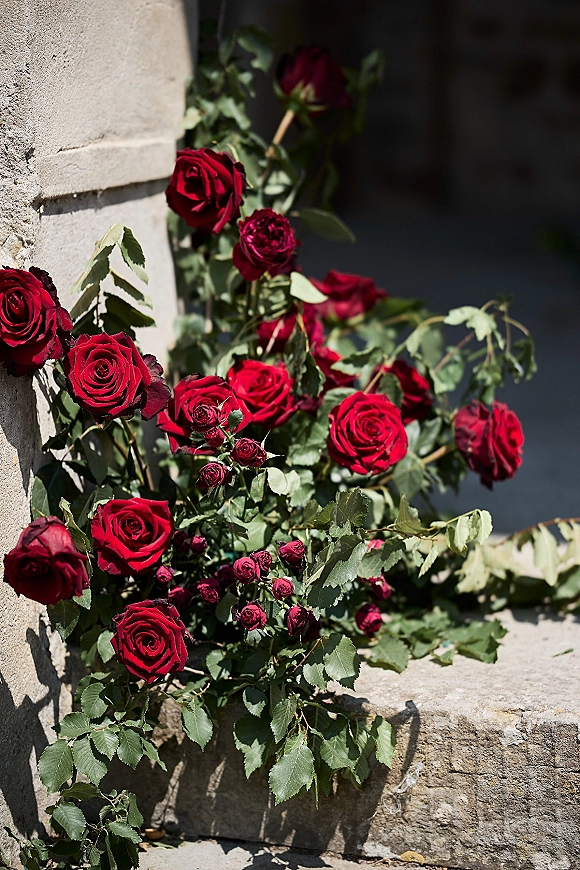 Rose arrangement of red roses and buds with greenery accents resting on a stone ledge against a textured stone wall in shadow