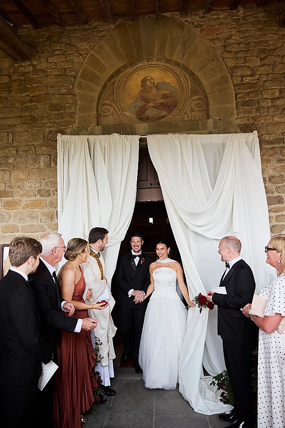 Wedding recessional as bride and groom exit a stone church doorway while guests toss rose petals under white draping