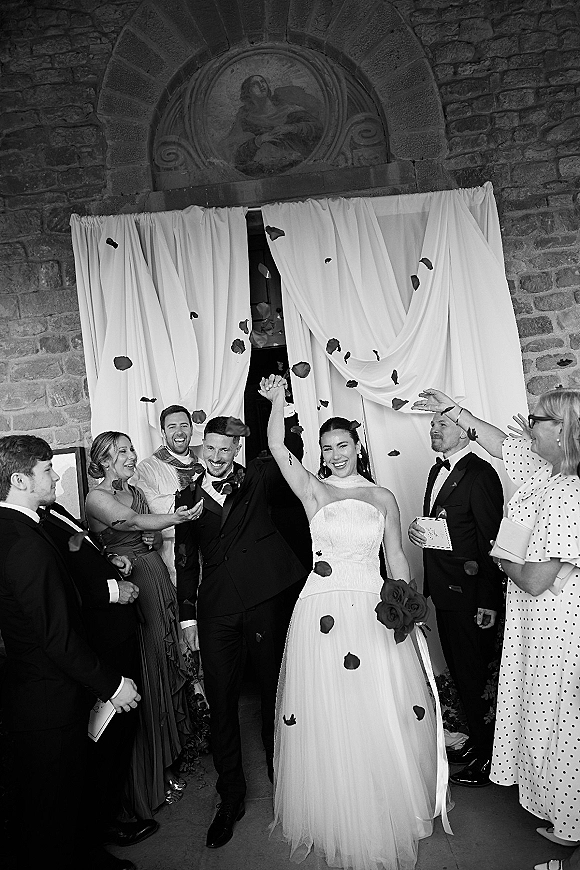 Wedding recessional as bride and groom exit beneath a stone church arch, rose petals tossed by cheering guests; bride raises arm holding bouquet