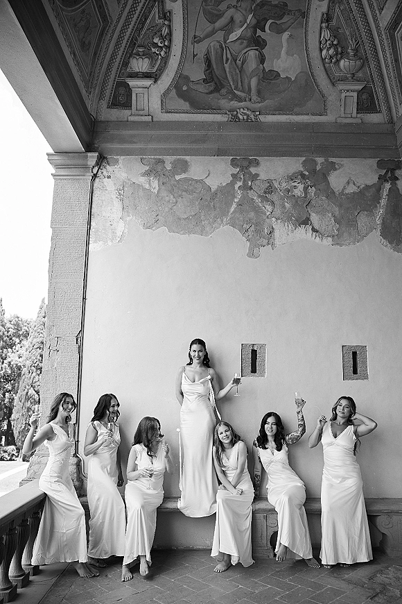 Bridesmaids portrait with champagne flutes, in matching satin slip gowns, posed under an ornate fresco ceiling on a balcony railing